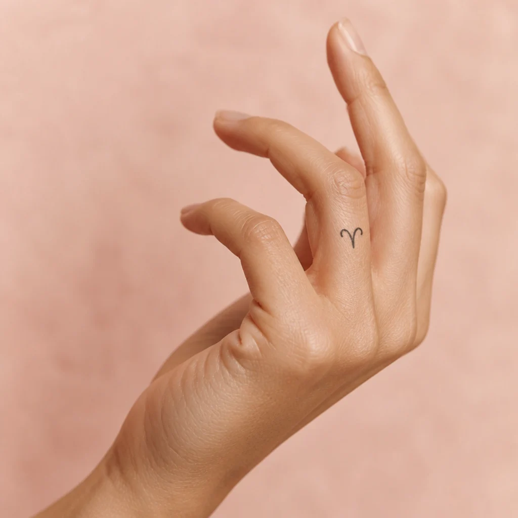 Tiny Aries glyph tattoo on the side of the finger in small black line on light-medium skin